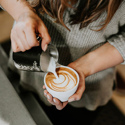 Eine Barista Frau die mit Milch und einer Kaffeetasse mit Kaffee drinnen und sie macht einen Cappuccino mit einem Muster drüber