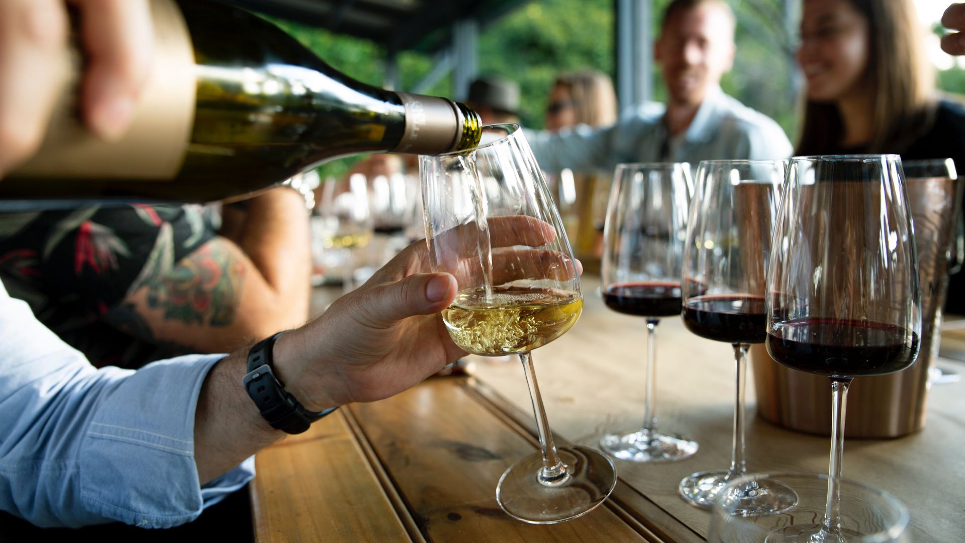 6 Menschen sitzen an einem Tisch bei einer Weinverkostung wo der Mann gerade den Weißwein einschenkt
